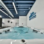 filled endless pools swim spa inside the lifestyle outdoor culver city showroom with churning water, resistance bars mounted across the pool, blue exposed ceiling beams above, black promotional balloons in the background, and the endless pools fitness systems wall logo on the right