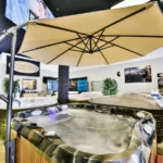 interior photo of the lifestyle outdoor sherman oaks showroom showing a filled hot tub with bubbling water beneath a large patio umbrella, with additional hot tubs, product signage, potted plants, and jacuzzi wall branding in the background.