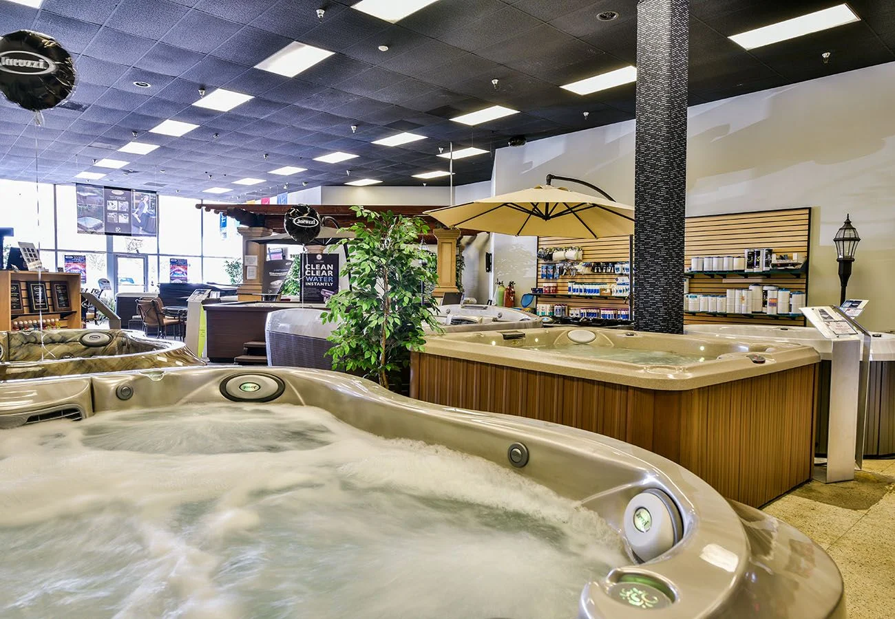 Interior photo of the Lifestyle Outdoor Sherman Oaks showroom showing a filled hot tub with bubbling water in the foreground, along with additional hot tubs, a spa supply wall, patio umbrella, potted plants, and showroom displays in the background.