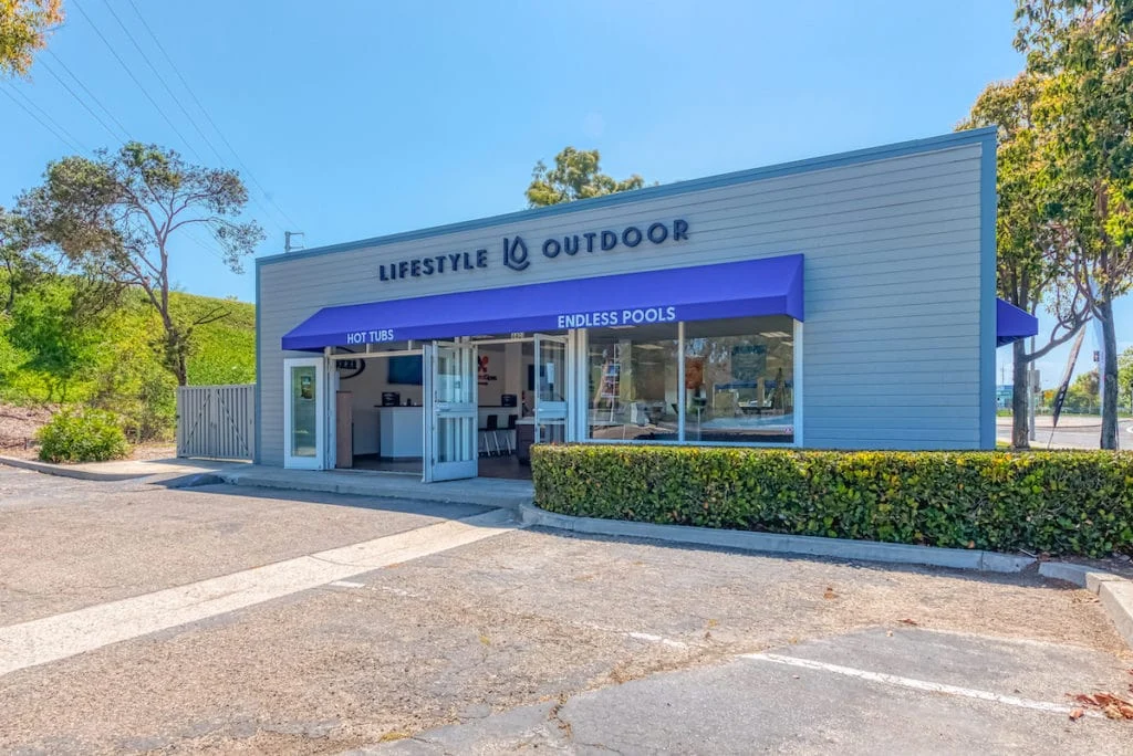 Front exterior photo of the Lifestyle Outdoor Ventura showroom showing the storefront sign, blue awning with Hot Tubs and Endless Pools text, open entrance doors, front windows, and parking area in front of the building.