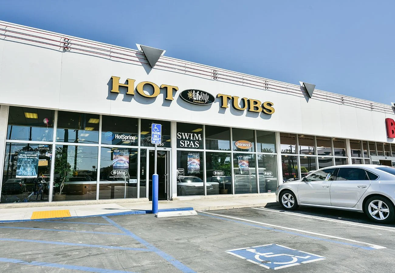 Front exterior photo of the Lifestyle Outdoor Sherman Oaks showroom featuring the main Hot Tubs storefront sign, Swim Spas window signage, brand logos on the windows, and the parking area in front of the entrance.
