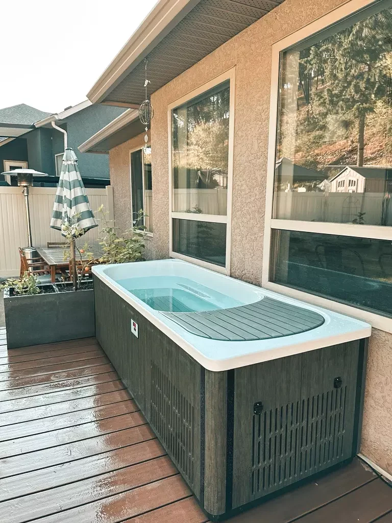 rectangular plunge tub with dark gray wood-look sides and a white rim on a wet backyard deck beside large house windows.