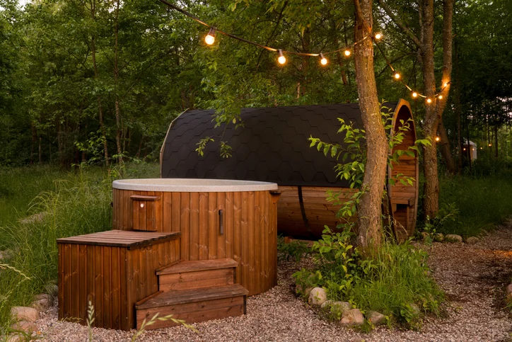 Wooden hot tub and barrel sauna set in a forest clearing at dusk, surrounded by tall trees, gravel paths, and warm string lights hanging overhead.